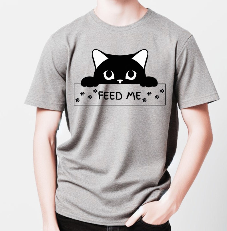 May include: A gray t-shirt with a black cat peeking over a sign that says "FEED ME". The sign has paw prints on either side.