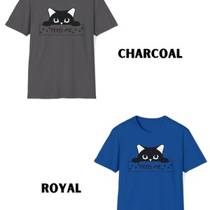 May include: A black cat peeking over a sign that says "Feed Me" with paw prints on either side. The sign is on a charcoal grey t-shirt. The same design is on a royal blue t-shirt.