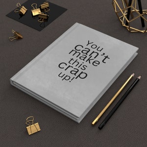 Puede incluir: Un diario gris claro con el texto "You can't make this crap up!" en negro. Lápices dorados y negros y clips dorados están colocados alrededor del diario sobre una superficie gris oscuro.