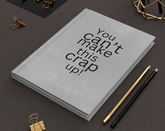 May include: A light gray journal with the text "You can't make this crap up!" in black. Gold and black pencils and gold binder clips are arranged around the journal on a dark gray surface.