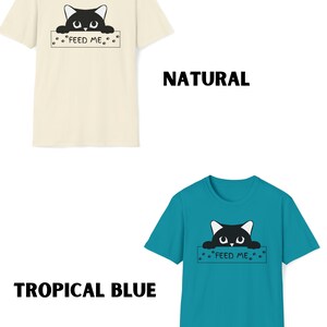 May include: Two t-shirts with a black cat peeking over a sign that says "Feed Me". The t-shirt on the left is a natural color and the t-shirt on the right is tropical blue.