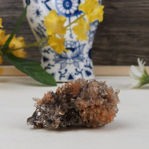 May include: A cluster of crystals with a mix of orange, brown, and clear points. The crystals are in focus, with a blurred background featuring yellow flowers, a blue and white patterned vase, and white flowers. The crystals are on a white surface.