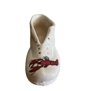 May include: A white ceramic boot-shaped vase with a red lobster illustration on the front. The vase has a V-shaped opening and small gold-colored accents. This decorative vase is suitable for home decor.