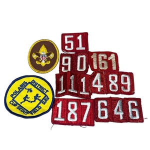 May include: A collection of vintage Boy Scout patches. Includes a round brown and gold patch with a fleur-de-lis emblem, a yellow and blue Polaris District patch, and several red patches with white numbers.