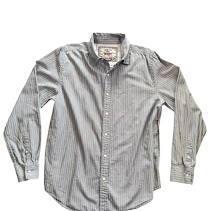 Puede incluir: Camisa de manga larga con rayas blancas verticales sobre fondo gris. La camisa tiene cuello clásico y puños abotonados. La etiqueta de la marca Levi's es visible en el cuello.