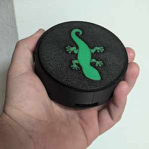以下が含まれることがあります： 黒い丸い容器で、表面はざらざらしており、上部に明るい緑色のトカゲのデザインがあります。トカゲは様式化されており、蓋の中央に配置されています。容器は手で持たれています。