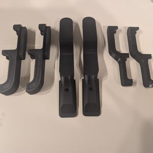 May include: Six dark grey 3D-printed plastic components are displayed. The parts include two long, flat pieces, two L-shaped pieces, and two smaller, angled pieces. The objects are arranged on a light-coloured surface.