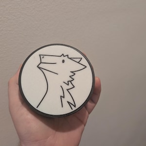 May include: A circular white coaster with a black edge, displaying a simple black line drawing of an animal head looking upwards. The design is set against a white backdrop.