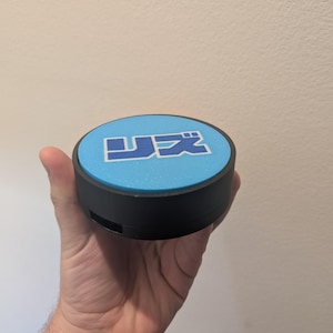 May include: A circular black object with a light blue top. The top displays Japanese characters in blue. The object is held in a hand against a plain background. The object appears to be a button or similar device.