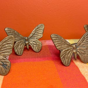 May include: Close-up of several bronze butterfly-shaped decorative objects. The butterflies are arranged in a row, with detailed wing patterns. The background is a vibrant orange wall and a colorful plaid fabric.