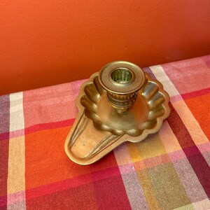 May include: A brass candlestick holder featuring a scalloped, circular base with a flared, extended handle. The central candle socket is cylindrical with a raised rim, and the entire piece has a polished, metallic gold-toned finish.