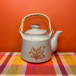 May include: A light blue ceramic teapot with a woven handle and a lid. The teapot features a wheat and butterfly design in shades of brown. The teapot is on a colorful plaid surface.