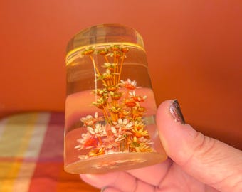 May include: A clear, cylindrical resin paperweight with dried flowers encased inside. The flowers are a mix of white, orange, and yellow, set against a transparent background. The paperweight is held in a hand.