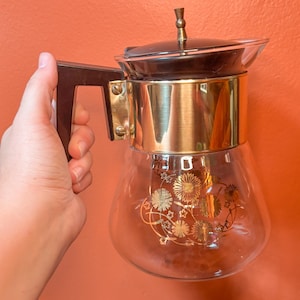 May include: A glass coffee pot with a gold band and a floral design. The pot has a brown plastic handle and a black lid.
