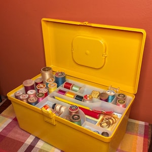 May include: A bright yellow plastic sewing box with an open lid, containing various colorful spools of thread, metal rings, and sewing notions organized in internal compartments. The box features a rectangular shape with a secure latch closure.