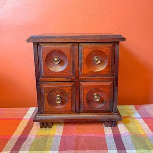May include: A small, wooden jewelry box with four drawers. The box is made of dark wood and has brass knobs. The drawers are square with a circular design in the center. The box sits on a plaid surface against an orange background.