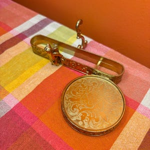 May include: A gold-toned compact mirror with an ornate floral design and a matching holder. The holder features a decorative cherub and scrollwork. The items rest on a colorful plaid fabric.