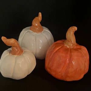 Puede incluir: Tres calabazas de cerámica, una naranja y dos blancas, con tallos marrones. Las calabazas son un artículo de decoración de otoño.