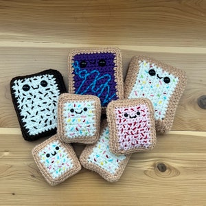 Puede incluir: Una colección de siete juguetes de peluche tejidos a crochet con forma de tartas pop. Los juguetes son de varios colores y tienen diferentes rellenos, incluyendo chocolate, fresa y arándano. Cada juguete tiene una cara sonriente con ojos negros y una boca blanca.