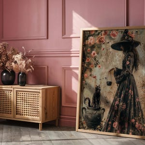 May include: A framed print of a woman in a black dress and witch's hat standing in a garden with a black cat. The woman is holding a cauldron and there are flowers and vines in the background.