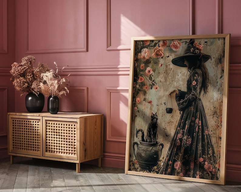 May include: A framed print of a woman in a black dress and witch's hat standing in a garden with a black cat. The woman is holding a cauldron and there are flowers and vines in the background.
