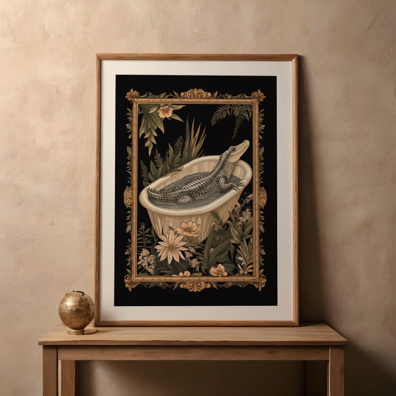 Op de afbeelding: Ingelijste kunstdruk met een alligator in een vintage badkuip. Het kunstwerk staat tegen een zwarte achtergrond met bloemen en botanische elementen, omlijst door goud. De print wordt op een houten tafel getoond.