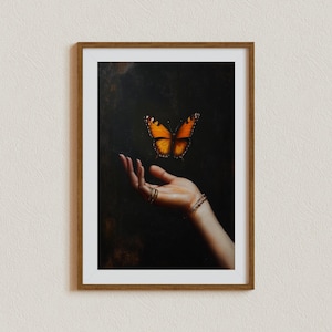 May include: Framed artwork featuring a vibrant orange and black butterfly hovering above an open hand. The hand is adorned with gold rings and bracelets, set against a dark background. The artwork is encased in a brown wooden frame.