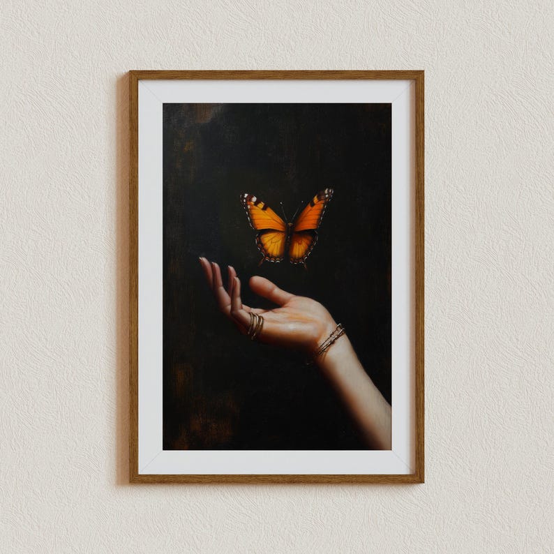 May include: Framed artwork featuring a vibrant orange and black butterfly hovering above an open hand. The hand is adorned with gold rings and bracelets, set against a dark background. The artwork is encased in a brown wooden frame.