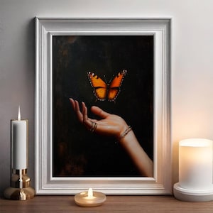 May include: A framed painting depicts a butterfly hovering above a hand adorned with rings and a bracelet. The butterfly has orange and black wings. The artwork is set against a dark background, framed in white, and displayed on a wooden surface.