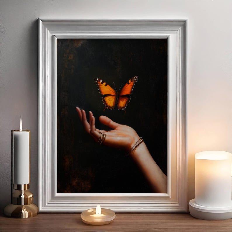 May include: A framed painting depicts a butterfly hovering above a hand adorned with rings and a bracelet. The butterfly has orange and black wings. The artwork is set against a dark background, framed in white, and displayed on a wooden surface.