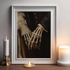 May include: A framed artwork showing two skeletal hands clasped together against a dark backdrop. The hands are intricately detailed, with visible bones. The white frame surrounds the artwork, which is displayed on a wooden surface with candles.