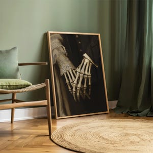 May include: A framed artwork showing skeletal hands clasped together, set against a dark backdrop. The hands are detailed, with visible bones. The artwork is in a wooden frame, leaning against a green wall. The image evokes themes of gothic art and Halloween.