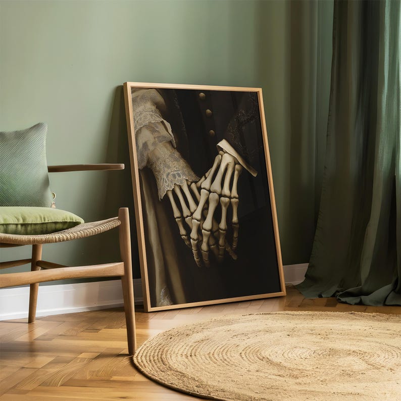 May include: A framed artwork showing skeletal hands clasped together, set against a dark backdrop. The hands are detailed, with visible bones. The artwork is in a wooden frame, leaning against a green wall. The image evokes themes of gothic art and Halloween.
