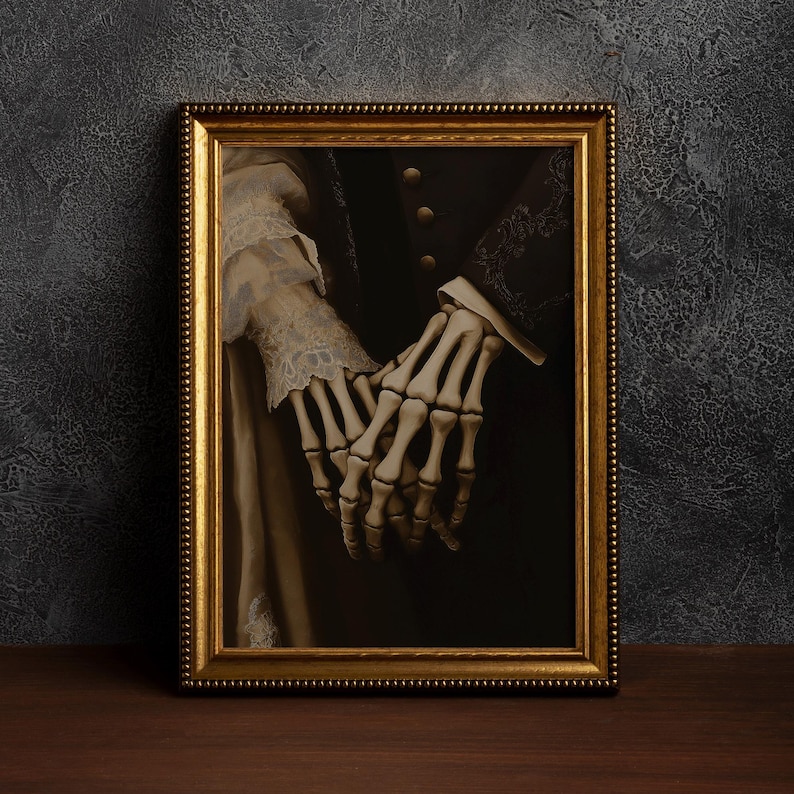 May include: A framed artwork showing a skeletal hand holding a human hand. The gold frame surrounds a dark, gothic image. The human hand is wearing a white lace sleeve, creating a contrast.