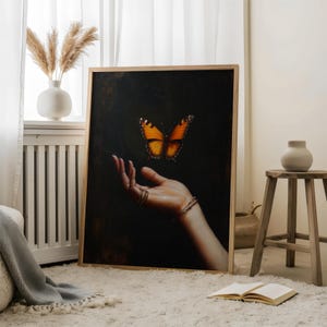 May include: A framed art print featuring a butterfly with orange and black wings hovering above a hand. The artwork is set against a dark background, with the hand adorned with rings and bracelets. The frame is made of light-colored wood.