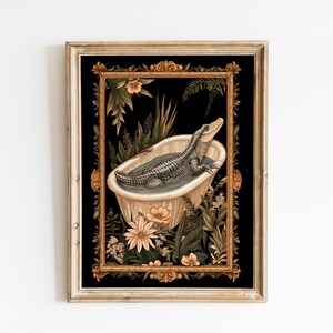 Op de afbeelding: Ingelijste kunstprint met een alligator die ontspant in een vintage badkuip. Het kunstwerk heeft een zwarte achtergrond met een gouden, sierlijke lijst en is omgeven door weelderig groen en bloemen. De alligator is gedetailleerd in grijstinten.