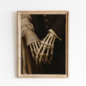 May include: A framed sepia-toned artwork depicting two skeletal hands holding each other. The hands are the central element, set against a dark backdrop. The artwork is enclosed in a gold-coloured, ornate frame, giving it a vintage appearance.