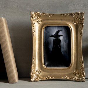 May include: A framed black and white illustration of a witch wearing a pointed hat and a long dress. The frame is gold and ornate.