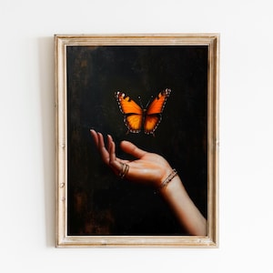 May include: A framed artwork featuring a hand with gold rings and bracelets, with an orange and black butterfly hovering above. The background is dark, and the frame is gold.