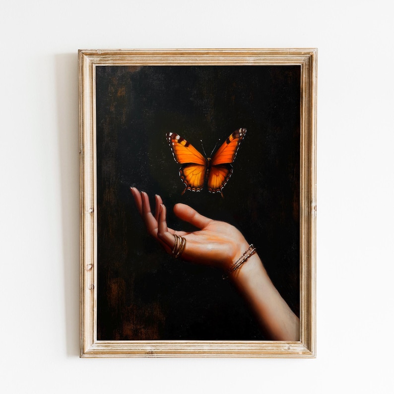 May include: A framed artwork featuring a hand with gold rings and bracelets, with an orange and black butterfly hovering above. The background is dark, and the frame is gold.