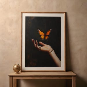 May include: A framed art print featuring a hand with multiple rings and bracelets, with an orange and black butterfly hovering above. The artwork is set against a dark background, with a wooden frame.