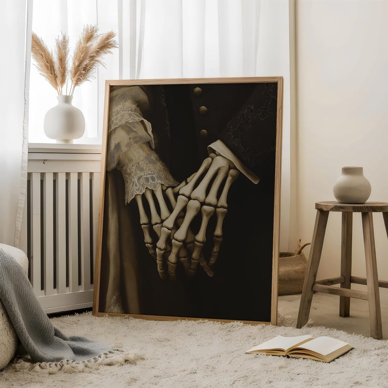 May include: A framed artwork depicting a skeletal hand holding a human hand, set against a dark backdrop. The human hand is decorated with lace, and the artwork is in a wooden frame. The overall theme is gothic and macabre.