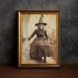 May include: A black and white vintage photograph of a woman dressed as a witch, wearing a pointed hat and a long dress. She is sitting on a rock in a forest setting.