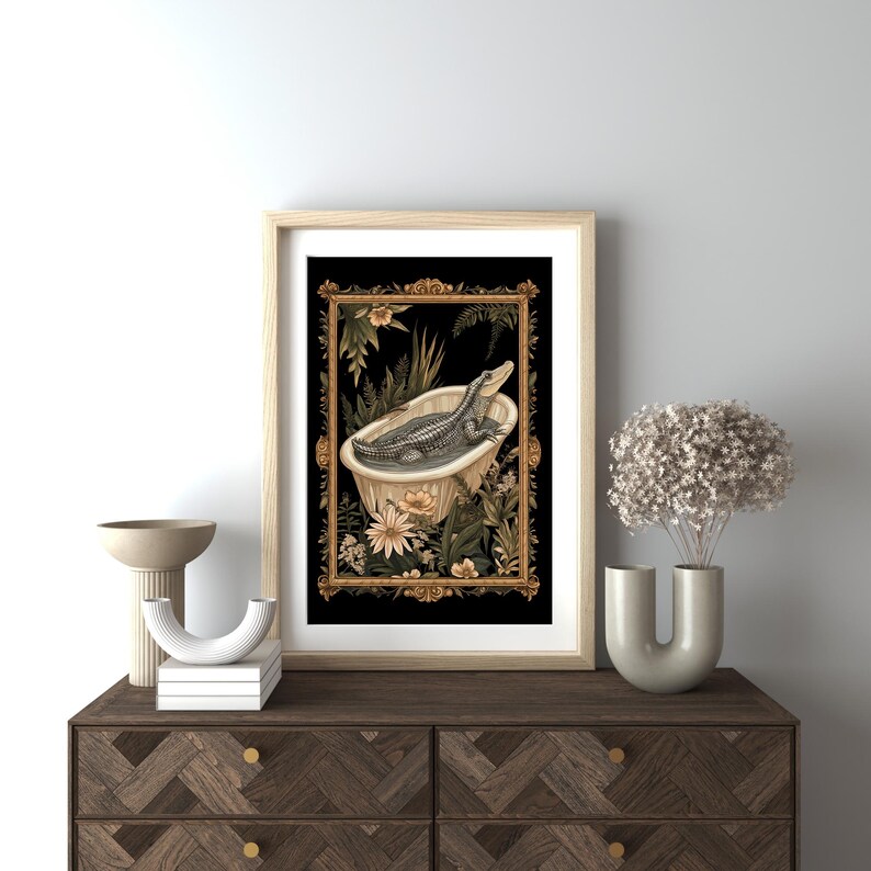 Op de afbeelding: Een ingelijste kunstdruk met een alligator in een badkuip, omringd door bloemen en botanische elementen. Het kunstwerk heeft een zwarte achtergrond en is ingelijst in een licht houten frame met gouden details.
