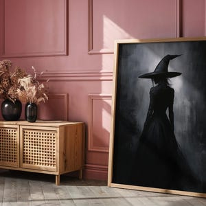 May include: A framed black and white illustration of a woman wearing a witch's hat and dress, standing in a foggy forest. The frame is made of wood and has a light brown finish.