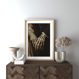 May include: A framed art print showing a sepia-toned image of two skeletal hands holding each other. The hands are the central element, set against a dark backdrop. The artwork is presented in a light wood frame, adding a touch of elegance.