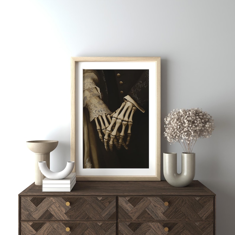 May include: A framed art print showing a sepia-toned image of two skeletal hands holding each other. The hands are the central element, set against a dark backdrop. The artwork is presented in a light wood frame, adding a touch of elegance.