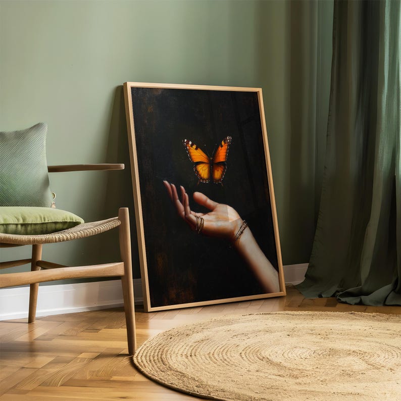 May include: A framed art print featuring a hand with rings and a bracelet, with a vibrant orange and black butterfly hovering above. The artwork is set against a dark background, with a wooden frame. The print is leaning against a wall.
