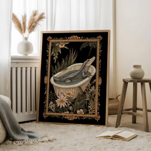 Op de afbeelding: Ingelijste kunstafdruk met een alligator in een vintage badkuip. Het kunstwerk staat tegen een zwarte achtergrond met weelderig groen en bloemen. De lijst is goudkleurig en rijk versierd, wat een vleugje elegantie toevoegt.
