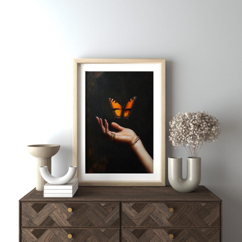 May include: Framed art print featuring a hand with rings and a bracelet, with an orange and black butterfly hovering above. The artwork is in a light wood frame, displayed on a dark wood dresser.